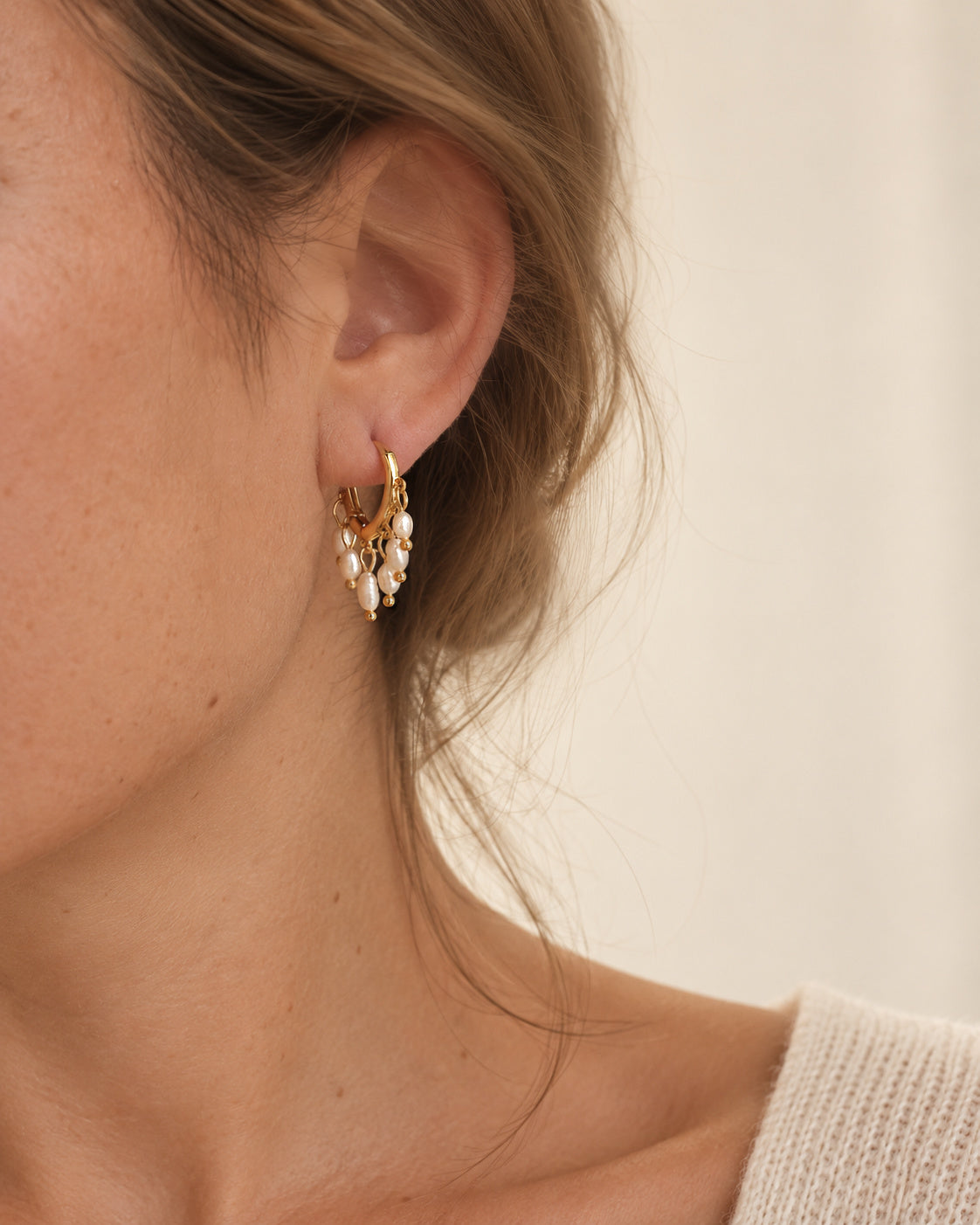 A close-up of small gold huggie hoop earring adorned with vintage white pearl fringe. Calitha Huggie Earring – White Pearl – A handcrafted earrings by Alysian.