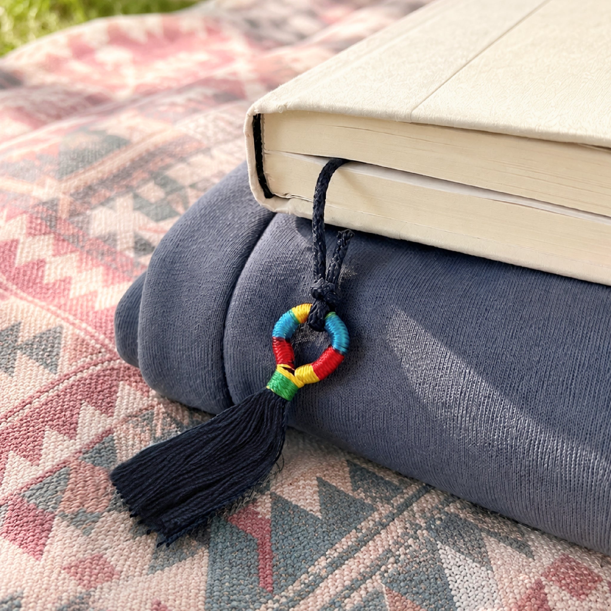 Colorful tassel Inanna Bookmark peeks out of book on top of a folded sweatshirt and a textural boho picnic blanket outdoors.
