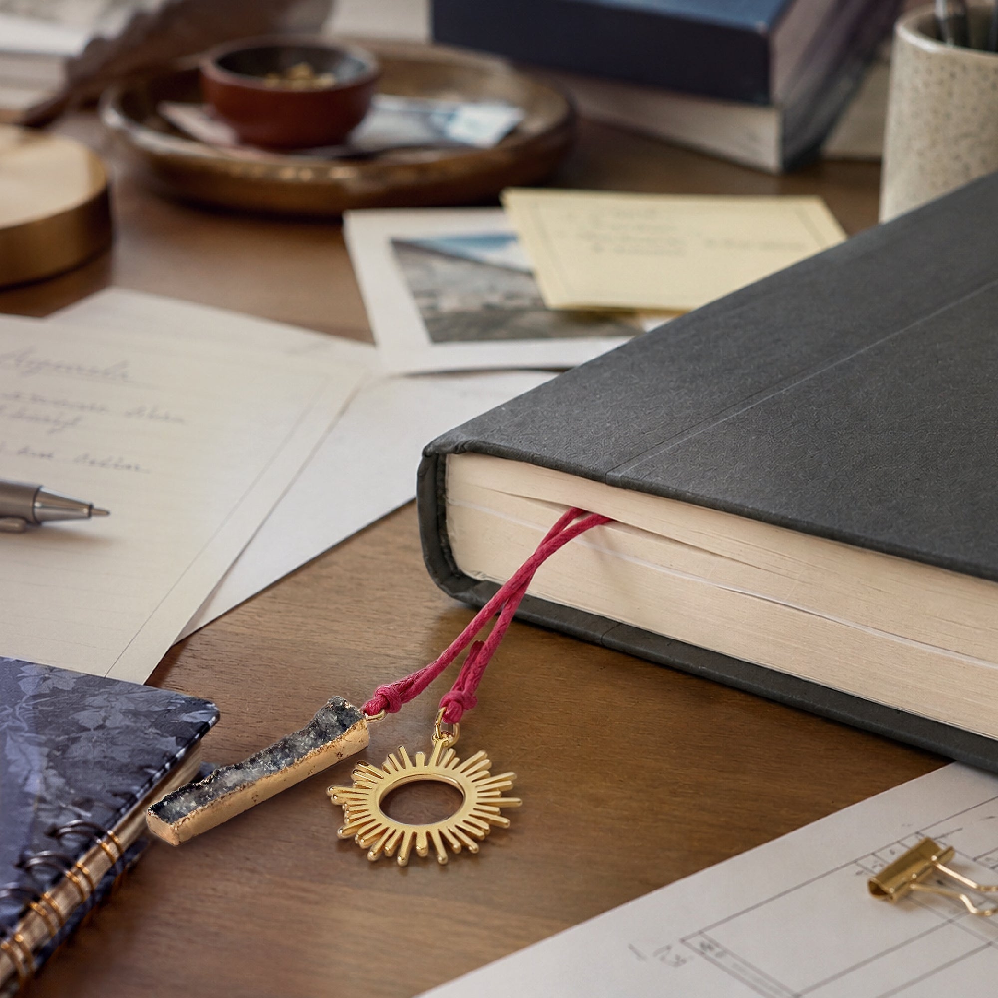 Nova bookmark with a gold sun-shaped pendant and rectangle gray druzy stone on fuschia cotton cord from a book with stationery items on a wooden desk.