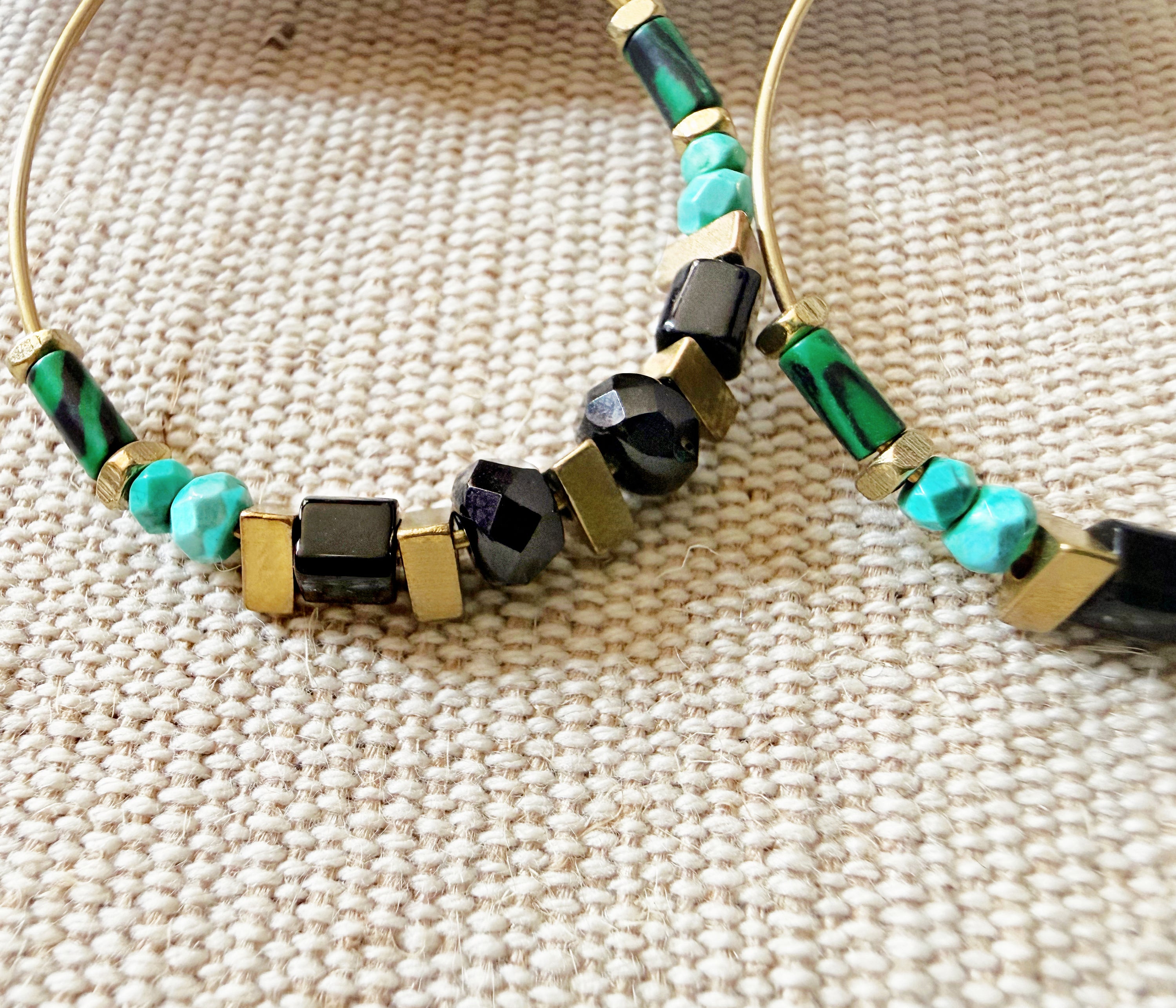 Closeup photo of the Adira Hoops, showing the details and textured of green malachite and faceted turquoise gemstones with black glass and brass geometric beads.