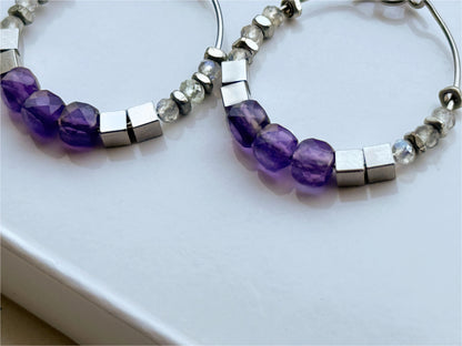 Detailed closeup of Luma Amethyst Hoop earrings with facets of amethyst beads, smoothness of stainless steel cube beads and shimmer of labradorite gemstones shown. 