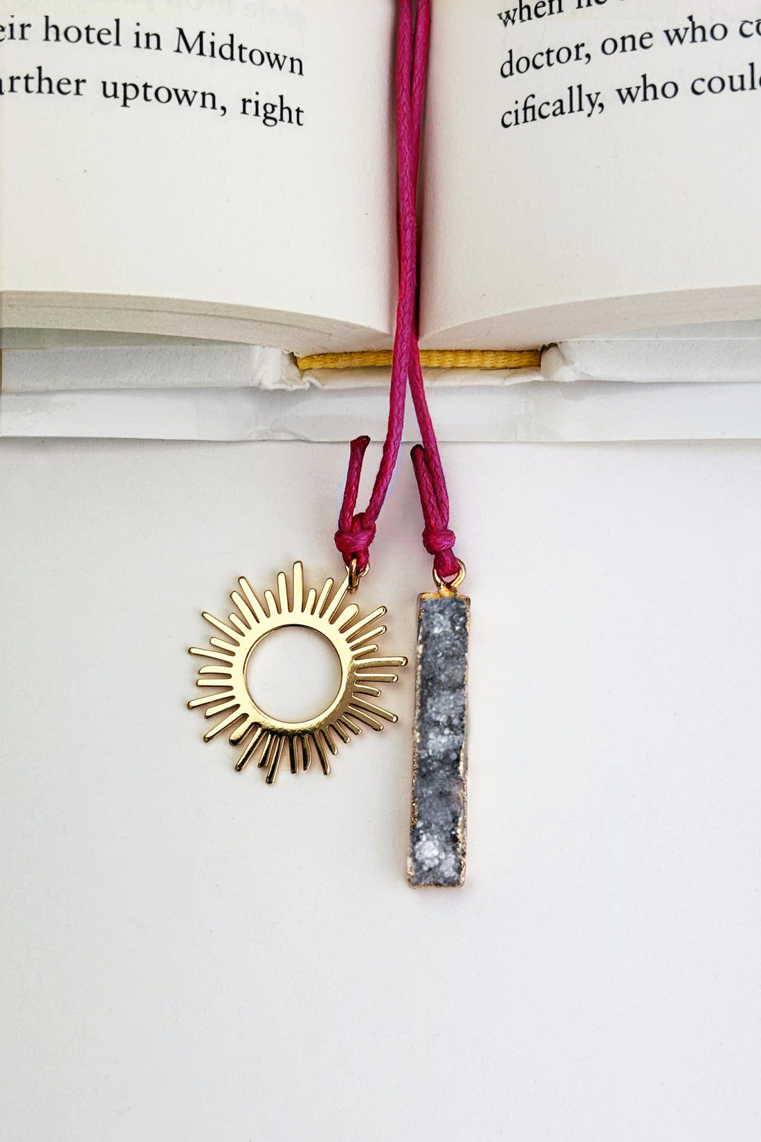 A gold sunburst pendant and a gray druzy stone bar pendant on a pink cord, displayed on an open book - Nova Druzy Bookmark – A handcrafted bookmark by Alysian.