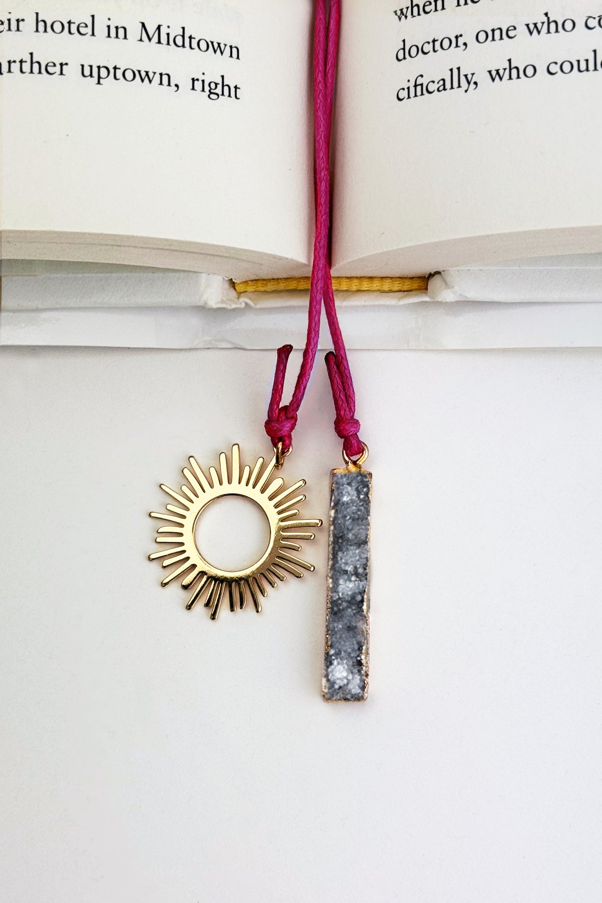 A gold sunburst pendant and a gray druzy stone bar pendant on a pink cord, displayed on an open book - Nova Druzy Bookmark – A handcrafted bookmark by Alysian.